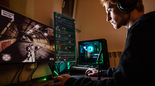 Na imagem temos um menino branco, cerca de 20 anos, cabelos curtos, com fones de ouvido e usando um moletom preto. Ele está sentado em frente a dois computadores, nos quais um deles tem uma imagem de jogo e o outro uma tela de programação.