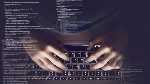 A imagem é uma foto: uma mão digital no teclado de um computador. Na tela do teclado, além dos códigos que estão sendo digitados, aparece o reflexo da mão
