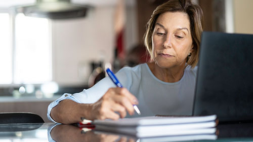 Na imagem uma mulher branca, de mais de 50 anos, cabelos curtos e lisos e vestindo uma camiseta branca, está sentada e segura uma caneta. Na mesa a sua frente está um computador e um caderno, onde ela escreve. A cena é ambientada em uma cozinha.