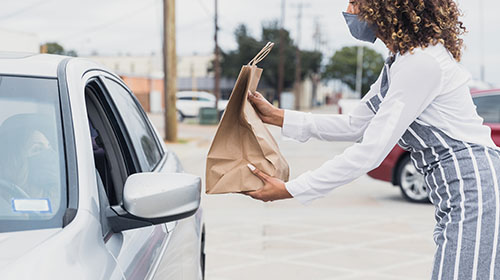 Na imagem uma mulher negra de cabelos cacheados, máscara, camiseta de manga longa branca e avental entrega uma sacola de papelão, pela janela, para outra mulher que está dentro de um carro.