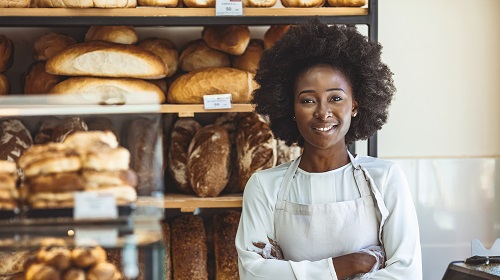 A imagem é uma foto de uma padaria. Há uma estante com pães diversos em cada prateleira ao fundo. Na frente, uma mulher negra de cabelos black power, camisa branca e avental branco, sorri.