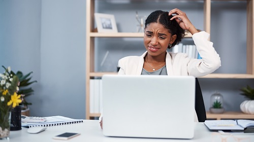 A imagem é uma foto de um escritório com uma estante de madeira ao fundo, com livros e porta-retratos. Uma mulher negra de cabelos presos, blusa azul e blazer branco está sentada em frente a uma mesa de trabalho. Ela está com uma expressão que indica confusão e coça a cabeça com a mão direita, olhando para um computador. Em cima da mesa estão alguns cadernos, uma calculadora e vasos de plantas.