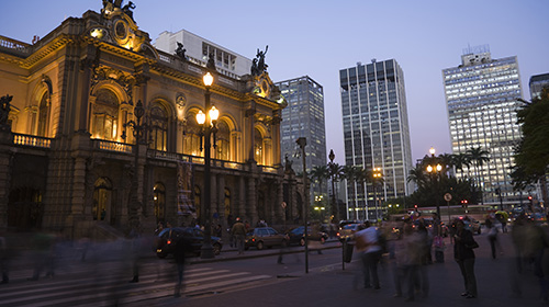 A imagem é uma foto. No centro de São Paulo está o prédio do Teatro Municipal e prédios mais modernos ao fundo. Pessoas caminham na rua.