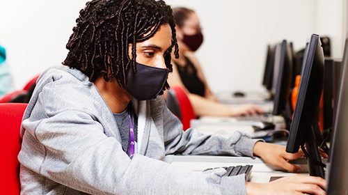 A imagem é uma foto. Um homem negro, jovem, está em uma sala de informática e olha para um computador