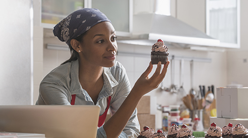A imagem é uma foto. Em uma cozinha, uma mulher negra, com uma bandana azul cobrindo os cabelos, camisa jeans e avental vermelho, apoia os cotovelos em uma mesa e segura com a mão direita um cupcake. Em cima da mesa, estão outros cupcakes de sabores variados e caixas de papel