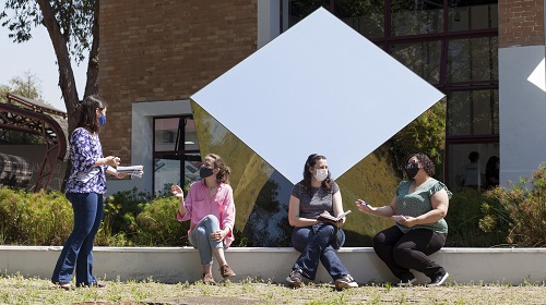 A foto é uma imagem. Em um ambiente externo de jardim, quatro mulheres estão conversando. Elas são brancas, usam máscaras de proteção contra a Covid-19 e duas delas seguram cadernos.