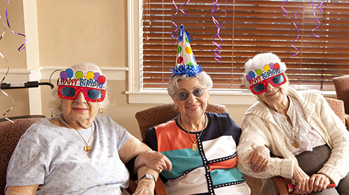 A imagem é uma foto de três senhoras idosas, sentadas lado a lado e com os braços entrelaçados. Elas estão com óculos e chapéus comemorativos em que está escrito "Feliz Aniversário".