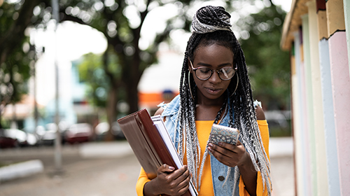 A imagem é uma foto. Uma jovem negra, de cabelos longos trançados e óculos arredondados, está parada na rua olhando para o celular. Ela carrega, em uma das mãos, um fichário e um caderno.