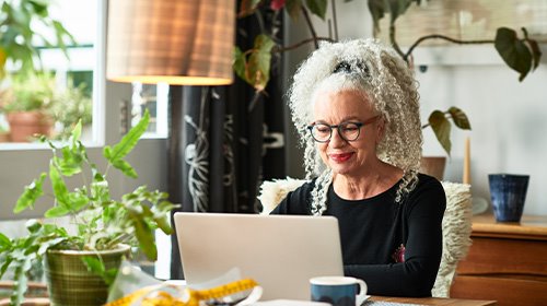 A imagem é uma foto. Uma senhora de cabelos brancos, cacheados e longos está sentada na sala de casa. Na mesa à sua frente está um computador.