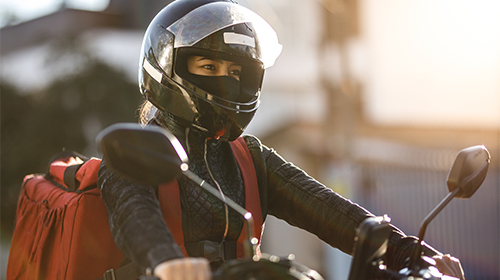 A imagem é uma foto. Uma mulher está em cima de uma moto. Ela está de capacete, jaqueta e uma mochila de entrega.