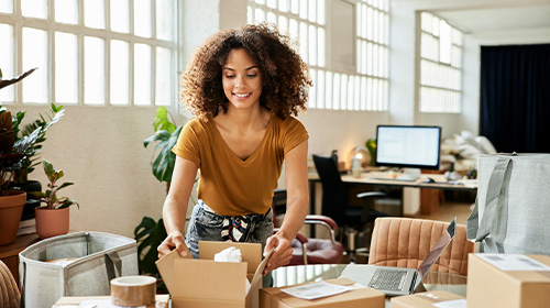 A imagem é uma foto em um ambiente de escritório, com mesas, cadeiras, computador e plantas. Uma mulher negra, de cabelos crespos longos, camiseta marrom e calça jeans, fecha caixas de papelão.  