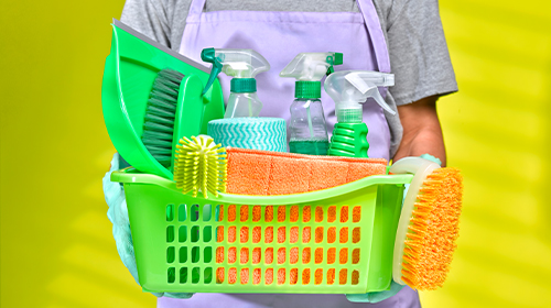 A imagem é uma foto. Uma pessoa de camiseta cinza e luvas roxas segura uma cesta verde. Dentro da cesta estão materiais e objetos utilizados na limpeza, como escovas, pá de lixo e produtos.