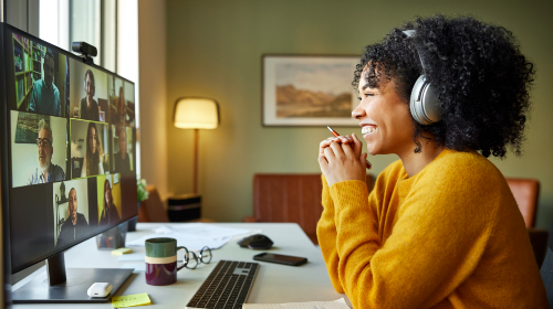 A imagem é uma foto de mulher negra, com cabelos crespos, fones e blusão amarelo, sentada em uma cadeira e de frente para uma tela de computador. Ela sorri e segura uma caneta. Na tela do computador aparecem rostos de outras pessoas.