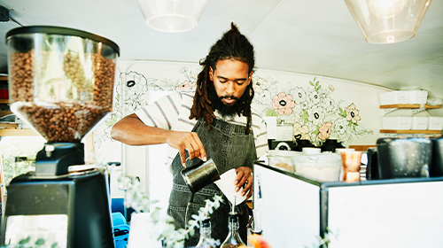 A imagem é uma foto. Em um ambiente de cafeteria, um homem negro, de dreads e avental jeans, está segurando uma xícara de metal com a mão esquerda e um copo de papel branco com a mão direita.
