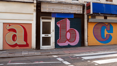 A imagem é uma foto. Na rua de uma cidade estão três lojas com as portas de metal fechadas. Em cada uma delas estão desenhadas em lettering as respectivas letras, a, b e c.