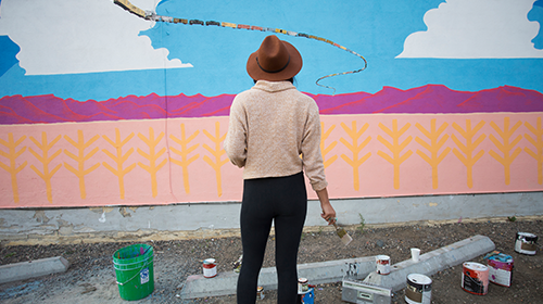 A foto é uma imagem. Um muro está pintado em tons de rosa, azul e branco, formando uma paisagem lúdica. Na calçada em frente ao muro estão latas de tinta e uma mulher, de costas para a câmera, segurando um pincel.