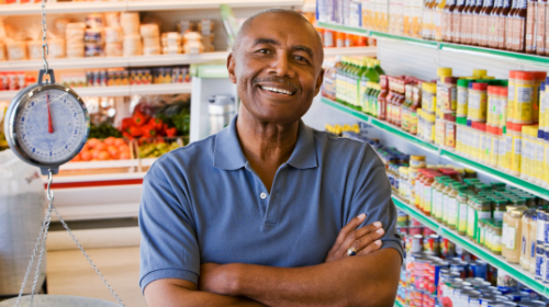 A imagem é uma foto. Em um supermercado, rodeado de prateleiras com produtos alimentícios, um homem negro, de camisa polo azul e avental branco, está sorrindo e de braços cruzados.