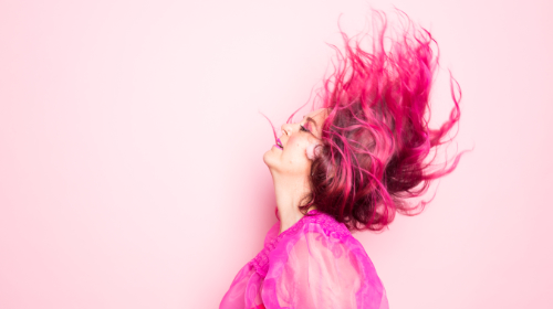 A imagem é uma foto. Em um fundo rosa, uma mulher de perfil joga os cabelos para o alto. Ela usa uma blusa de tule rosa e seu cabelo está tingido de rosa.