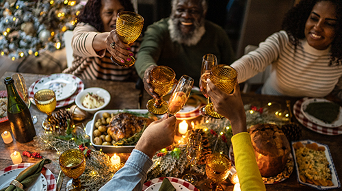 A imagem é uma foto. Cinco pessoas negras estão sentadas em cadeiras em volta de uma mesa. Elas levantam taças, fazendo um brinde. Em cima na mesa estão diversos pratos de comida e uma decoração natalina.