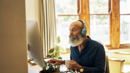 A imagem é uma foto. Em um ambiente de escritório, um homem negro está sentado e vendo algo no computador a sua frente.