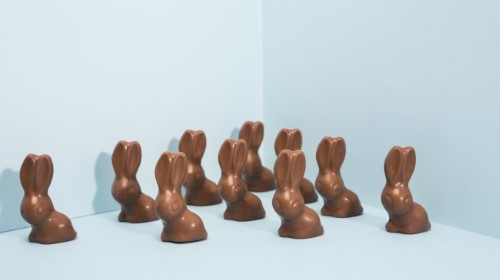 A imagem é uma foto. Em um fundo azul, coelhinhos de chocolate estão posicionados lado a lado.