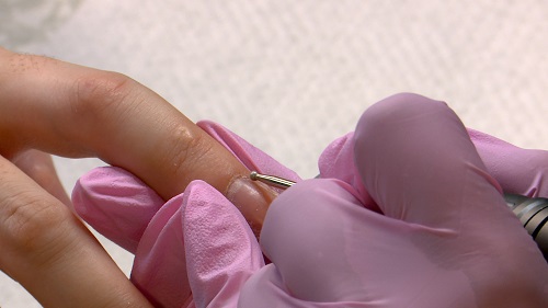 A imagem é uma foto. Mãos com luvas descartáveis na cor rosa estão segurando a mão de uma cliente e uma broca própria para unha, na linha da cutícula.