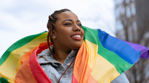 A imagem é uma foto. Uma mulher negra, de tranças e jaqueta jeans, está abraçada com uma bandeira da causa LGBTQIA+