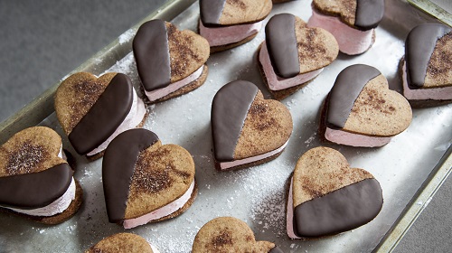 A imagem é uma foto. Uma assadeira contém diversos biscoitos em formato de coração, cobertos com chocolate e recheados com um creme cor de rosa.
