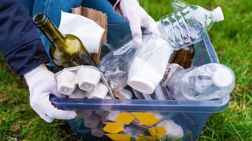 A imagem é uma foto. Dentro de um balde azul, estão diversos materiais recicláveis, como garrafas plásticas, garrafas de vidro e caixas de ovos de papel. Uma pessoa de luvas segura a caixa.