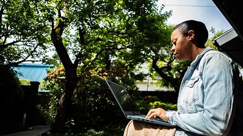A imagem é uma foto. Em um ambiente aberto e arborizado, uma mulher negra, de cabelos raspados, jaqueta jeans e saia bege, está sentada com um notebook em seu colo.