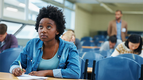 A imagem é uma foto. O ambiente é uma sala de aula, com carteiras azuis espalhadas e alguns alunos sentados fazendo anotações. O foco da imagem está em uma mulher de jaqueta e camisa azul olhando para a frente e segurando uma folha e uma caneta sobre a mesa.