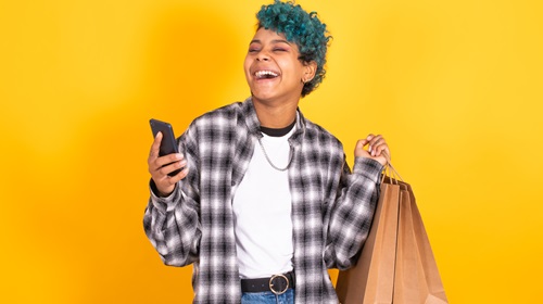 Mulher branca, de cabelos curtos na cor verde. Ela sorri, usa colar prata, calça jeans e cinto preto, blusa branca com gola preta e, por cima, camiseta de manga longa xadrez nas cores branco, cinza e preto. Na mão direita ela segura um celular preto e, na mão esquerda, sacolas de papel marrom-claras. As duas mãos estão na altura dos ombros.