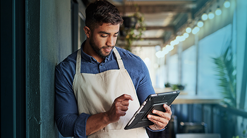 Na imagem, há um homem com barba, de pele morena, vestindo uma camisa azul e um avental branco e utilizando um tablet. Ele está encostado em uma parede de um corredor iluminado por luzes de teto, ao fundo.