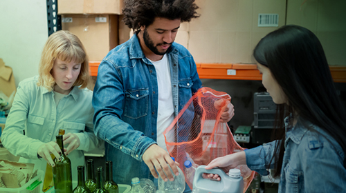 A imagem é a foto de três pessoas organizando materiais recicláveis. À esquerda, uma mulher de cabelo loiro curto e pele branca coloca garrafas de vidro em uma caixa plástica transparente com o símbolo de reciclagem. No centro, um homem de cabelo crespo volumoso, barba e pele preta vestido com uma jaqueta jeans, segura uma rede laranja com garrafas plásticas. À direita, uma mulher de cabelo preto longo e liso, pele branca vestida com uma jaqueta jeans, entrega mais garrafas ao homem. Em cima da mesa, há caixas de plástico com símbolos de reciclagem, uma contém garrafas de vidro e outra contém garrafas plásticas. Ao fundo, há estantes com caixas de papelão e outros itens armazenados.