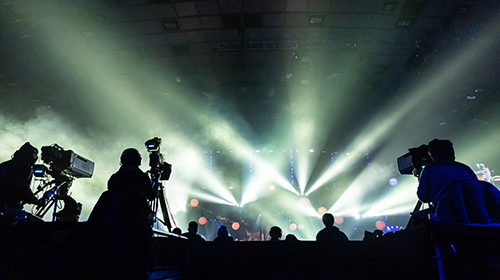 A imagem é uma foto. O ambiente tem pouca iluminação. Ao fundo, é possível ver um palco com jogo de luzes brancas refletidas. Mais próximo à câmera, vemos a silhueta de três equipamentos de filmagem e pessoas manuseando os equipamentos.