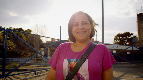 A imagem é uma foto. Em um ambiente a céu aberto, vemos ao fundo grades azuis, árvores e o céu com poucas nuvens. No centro da imagem, está uma mulher adulta, com cabelo loiro e curto, usando batom rosa e que veste uma camiseta roxa com estampa e brincos. Ela carrega uma bolsa apoiada no sentido transversal do seu corpo. Está sorrindo para a foto.