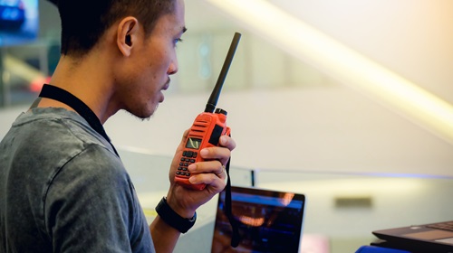 A imagem é uma foto. Um homem asiático de perfil segura um walkie-talkie. Em frente a ele, há equipamentos de som e dois notebooks.