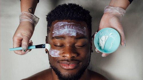 A imagem é uma foto. O foco está no rosto de um homem negro. Ele tem barba e cabelo curtos e está deitado sem camisa, com os olhos fechados e um sorriso. Duas mãos, uma de cada lado de sua cabeça, aparecem com luvas. A mão esquerda, à direita na foto, segura um pote azul com creme, enquanto a mão direita, à esquerda na foto, passa um pincel com o creme no rosto do homem.