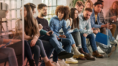 A imagem é a foto de um grupo de jovens adultos, diversos em aparência, sentado lado a lado em uma sala ampla e iluminada, em uma conversa informal. Vários olham uns para os outros enquanto sorriem, conversam ou escutam.