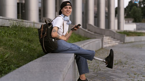 A imagem é uma foto. Em um ambiente externo, uma pessoa está sentada em uma mureta. Ela está de boné, fones de ouvido, camisa branca e uma calça jeans clara. Ela está com uma mochila nas costas, segurando um celular e uma de suas pernas é mecânica.